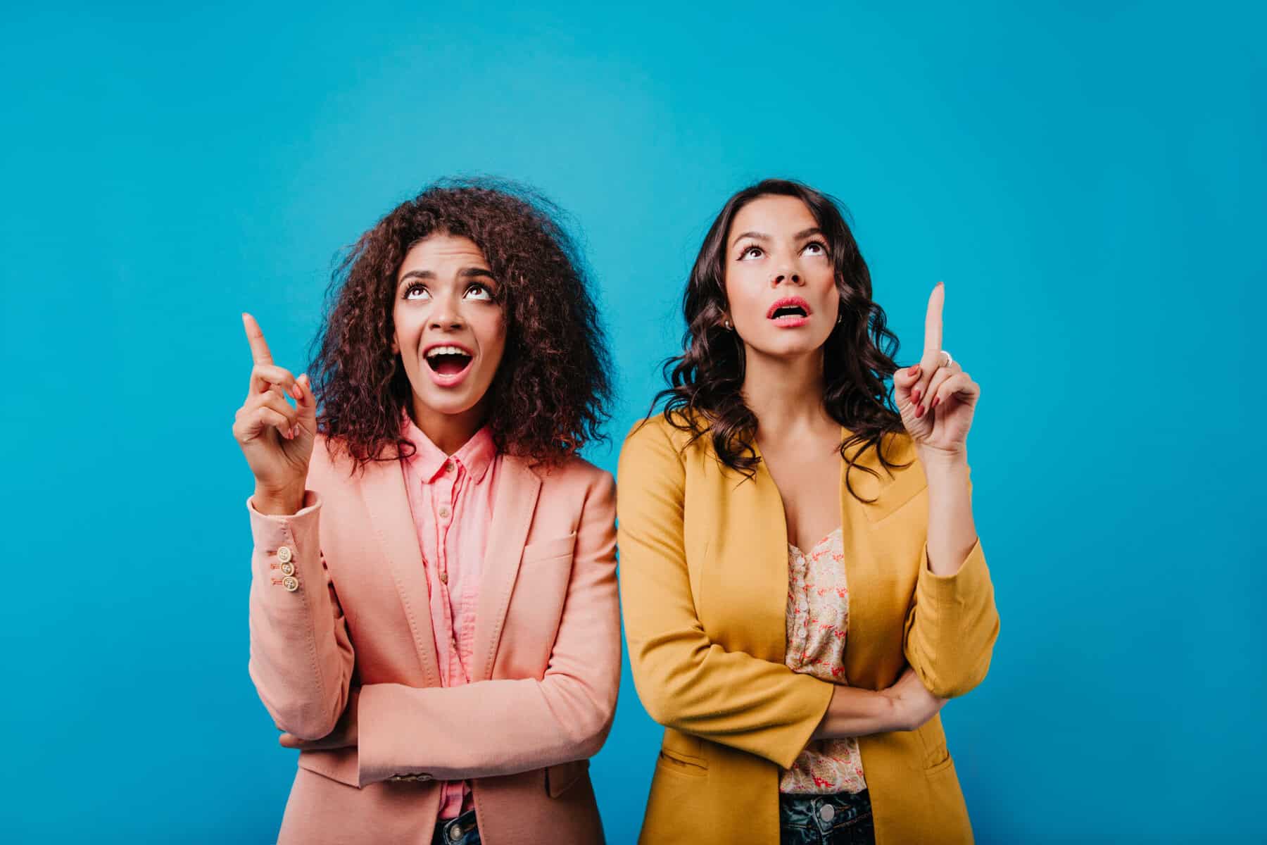 Deux femmes en veste colorée pointant du doigt vers le haut avec des expressions de surprise sur un fond bleu, illustrant des idées innovantes en gestion du risque de change pour 2025.